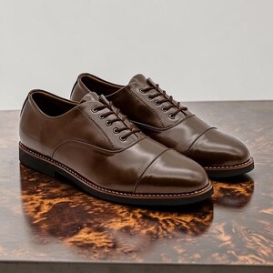 May include: A pair of brown leather dress shoes with a cap toe design. The shoes have a glossy finish, brown laces, and a black sole with a stitched border. The shoes are displayed on a wooden surface.