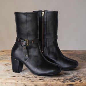 May include: A pair of black leather mid-calf boots featuring a block heel, a side zipper with a metallic pull, and a decorative strap across the ankle secured with a brass-toned buckle and eyelets. The boots have a smooth finish and rounded toes.