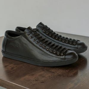 May include: A pair of low-top sneakers crafted from black leather, featuring matching black laces, silver-toned metal eyelets, and a solid black rubber sole. The side of the shoe displays a subtle embossed logo detail near the lacing area.