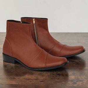May include: A pair of ankle-high boots in a smooth tan leather finish. These boots feature a rounded toe, a vertical gold-toned zipper on the side, and a low black heel. The design includes a stitched seam across the vamp and a plain shaft.