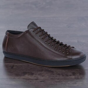 May include: A low-top sneaker in dark brown leather featuring a rounded toe, matching brown laces, and a solid black rubber sole. The side of the shoe displays a small embossed circular logo near the heel, with a clean, minimalist profile.
