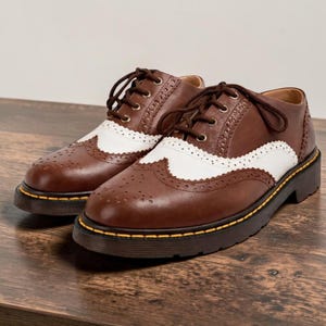May include: A pair of brown and white oxford shoes with a lace-up design. The shoes have a perforated pattern and a thick, black sole with a yellow trim. The shoes are made of leather and are displayed on a wooden surface.