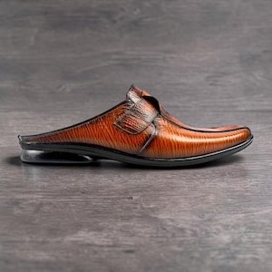 May include: A brown leather mule shoe with a textured finish. The shoe features a strap across the top and a black sole. The shoe is designed with a pointed toe and a low heel.