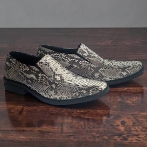 Pode incluir: Um par de sapatos sem atacadores com um padrão de pele de cobra cinzento e preto. Os sapatos têm um design de biqueira pontiaguda, um colarinho baixo e uma sola preta sólida. O material texturizado cobre toda a parte superior.