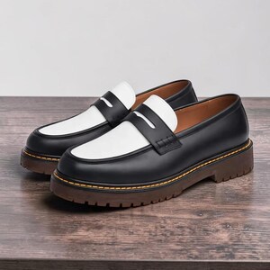 May include: Pair of black and white leather loafers with a chunky brown sole. The shoes have a classic penny loafer design with a white vamp and a black upper. The soles are approximately 1.5 inches thick and feature yellow stitching.