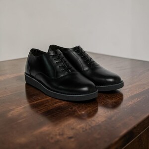 May include: A pair of black leather dress shoes with a low profile sole. The shoes are laced up and sit on a dark wooden surface. The shoes are a classic style, suitable for formal occasions.