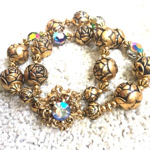 May include: Gold-toned beaded bracelet featuring rose-shaped beads and iridescent crystal accents. The bracelet includes a central decorative element with a large, iridescent crystal. The beads are approximately 0.75 inches in diameter.