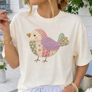 Puede incluir: Una camiseta de manga corta color marfil con un gráfico central de un pájaro compuesto por varios patrones de tela tipo patchwork con flores y cuadros en tonos rosa, morado, verde y amarillo, con un pequeño pico naranja y patas marrones.