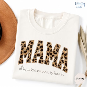 Puede incluir: Una camiseta de cuello redondo color marfil con la palabra MAMA en letras grandes de estilo bloque con un estampado de leopardo marrón y negro. Debajo de la palabra, los nombres olivia, emma y liam están impresos en una fuente cursiva negra.