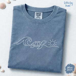 Puede incluir: Una camiseta de color azul vaquero con un gráfico de líneas blancas de una tortuga marina nadando cerca de una ola y un sol. La camiseta tiene cuello redondo y una etiqueta de la marca Comfort Colors en el interior. El tejido tiene un aspecto lavado.