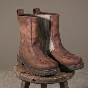May include: A pair of mid-calf brown leather boots featuring a rounded toe, a vertical seam detail along the front, and a thick, lugged rubber sole. The boots have a light-colored interior lining visible at the top and a sturdy construction.