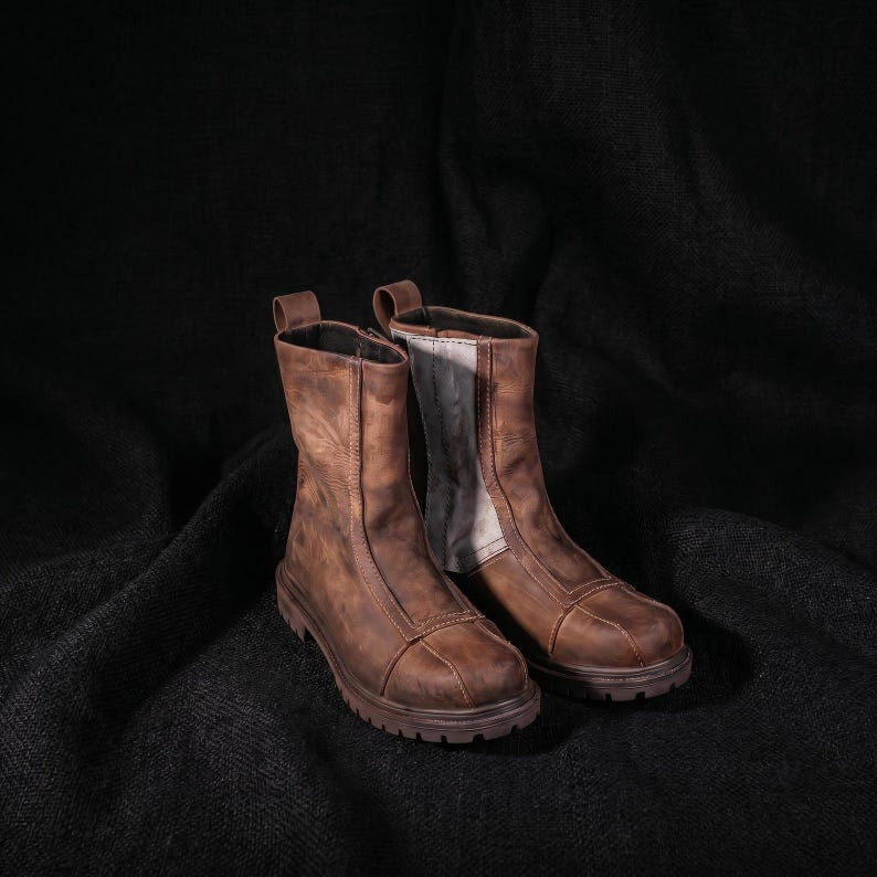May include: A pair of mid-calf brown leather boots featuring a distressed finish, a rounded toe cap with reinforced stitching, a vertical seam running up the front, and a thick, lugged rubber sole. The interior lining is visible in a light grey tone.