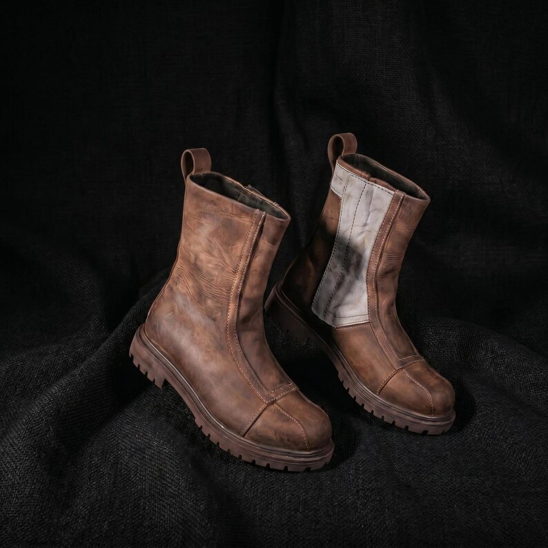 May include: A pair of mid-calf boots crafted from distressed brown leather. The boots feature a rounded toe, a thick lugged rubber sole, and a vertical pull tab at the back. One boot displays a contrasting light gray panel on the side shaft.