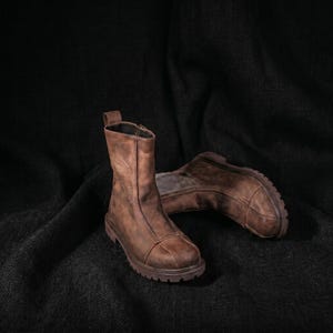 May include: A pair of mid-calf brown leather boots featuring a distressed finish, a vertical seam running down the front, a rounded toe cap, and a thick, lugged rubber sole. The boots have a pull tab at the top and a structured, sturdy design.