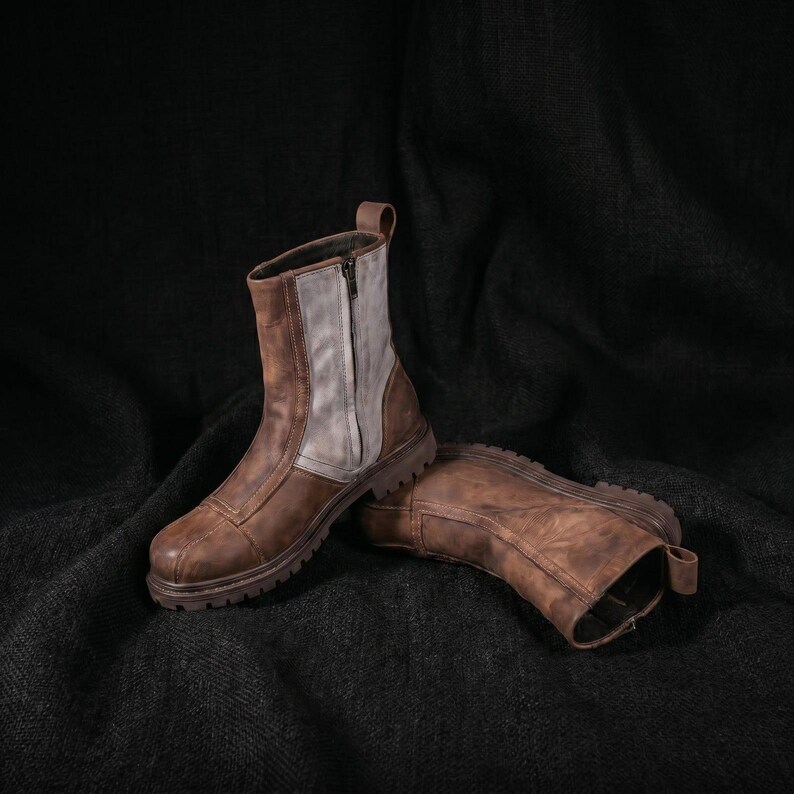 May include: A pair of mid-calf boots featuring a distressed brown leather construction with a contrasting light gray leather panel on the side. The boots include a vertical side zipper, a reinforced toe cap, and a thick, lugged rubber sole.