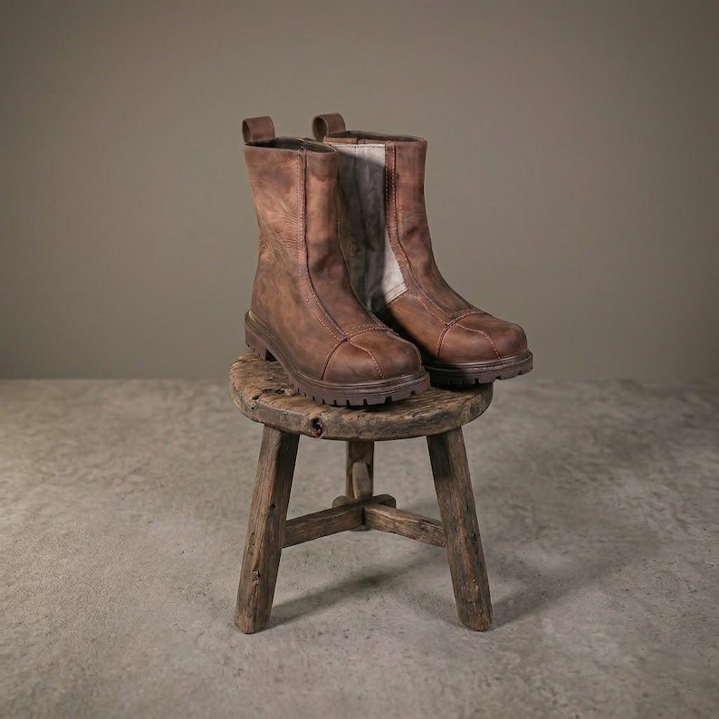May include: A pair of mid-calf brown leather boots featuring a rounded toe, a vertical seam detail along the front, and a thick, lugged rubber sole. The boots have a light-colored interior lining visible at the top and a sturdy construction.