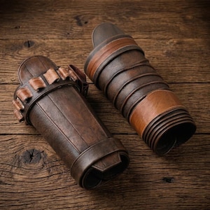 Puede incluir: Un par de protectores de brazo de cuero marr&oacute;n oscuro con un acabado texturizado. Un protector incluye una serie de peque&ntilde;os bucles a lo largo del borde superior, mientras que el otro presenta una banda de cuero marr&oacute;n claro lisa en el centro.
