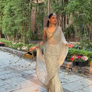 May include: A cream-coloured sari with gold embroidery and embellishments. The sari is draped over a gold-embroidered top. The woman is holding a gold handbag and wearing gold jewellery, including earrings and a headpiece. The background features bamboo trees and a stone walkway.