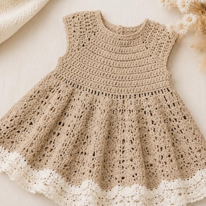 K&ouml;nnte beinhalten: Beige geh&auml;keltes Kleid mit &auml;rmellosem Design und ausgestelltem Rock. Das Kleid hat einen wei&szlig;en, gewellten Spitzenbesatz am Saum. Die H&auml;kelarbeit ist detailliert und zeigt ein strukturiertes Muster auf dem gesamten Kleidungsst&uuml;ck.