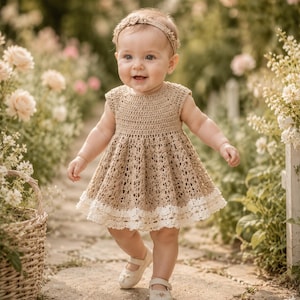 K&ouml;nnte beinhalten: Ein l&auml;chelndes Baby in einem hellbraunen H&auml;kelkleid mit wei&szlig;er Spitzenborte, passendem Stirnband und cremefarbenen Schuhen. Das Baby geht auf einem Steinweg in einem Blumengarten mit einem Weidenkorb.
