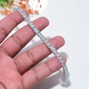 Op de afbeelding: Een zilveren armband met een rij lichtblauwe edelstenen. De armband wordt gepresenteerd tegen een lichte achtergrond, waardoor de glans van de stenen wordt benadrukt. Het sieraad is in klassieke tennisarmband-stijl.