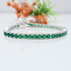 May include: A silver-toned tennis bracelet featuring a continuous row of round, vibrant green stones set in individual metal prongs. The bracelet has a secure clasp mechanism and a polished finish, creating a uniform and circular design.