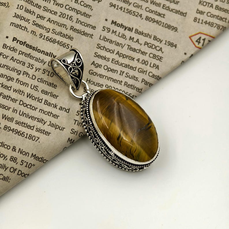 May include: An oval tiger's eye gemstone pendant set in a decorative silver frame with intricate metalwork patterns. The pendant features a polished, golden-brown stone with natural chatoyant bands and a matching silver bail at the top.