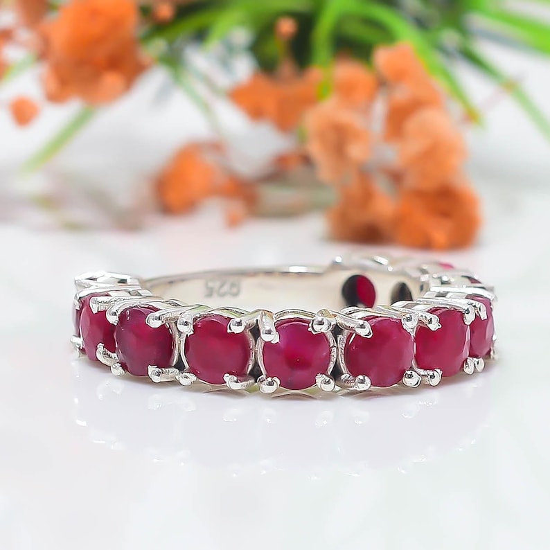 May include: A silver ring featuring a row of round, deep red gemstones set in individual metal prongs. The inner band of the ring is clearly stamped with the number 925, indicating the metal composition. The design is a continuous eternity style.
