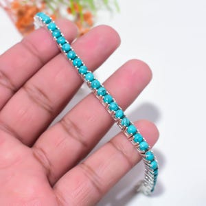 May include: A silver-toned tennis bracelet featuring a single row of round, bright turquoise beads with dark matrix veining. The beads are set in a metallic frame with individual prongs securing each stone along the length of the flexible bracelet.