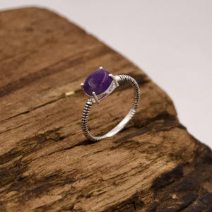 Könnte beinhalten: Ein Silberring mit einem glatten, ovalen violetten Edelstein in einer Vier-Krappen-Fassung. Die Ringschiene weist ein markantes, strukturiertes Design auf, das aus geripptem oder gewickeltem Draht besteht und den gesamten Ring umschließt.
