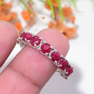 May include: A silver eternity band ring featuring a row of round, deep red faceted gemstones set in individual metal prongs. The ring displays a continuous line of stones across the top, showcasing a polished metallic finish and vibrant red colour.