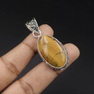 May include: An oval tiger's eye gemstone pendant set in a decorative silver frame. The stone features golden-brown tones with dark, chatoyant streaks. The silver bail at the top displays an intricate, openwork scroll design with small bead accents.