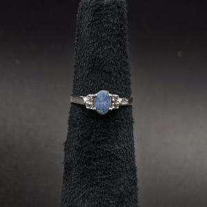 Könnte beinhalten: Ein silberner Ring mit einem ovalen, blauen Opal-Mittelstein. Der Ring hat kleine Akzentsteine auf beiden Seiten des Opals. Präsentiert auf einem schwarzen Samtringhalter vor dunklem Hintergrund.