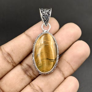 May include: An oval-shaped tiger's eye gemstone pendant set in a silver-toned metal frame. The stone features golden-brown bands with a chatoyant luster. The pendant includes a decorative silver-toned bail with intricate scrollwork detailing.