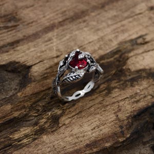 May include: A silver ring with a round, deep red gemstone at the center. The ring features a leaf design around the gemstone and along the band. The ring is set on a wooden surface.