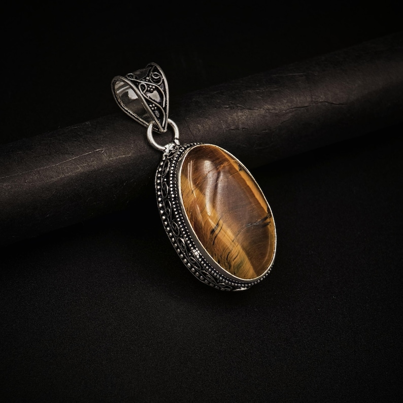 May include: An oval tiger's eye gemstone pendant set in a decorative silver frame with intricate metalwork detailing. The pendant features a polished, golden-brown stone with natural chatoyant bands and a matching silver bail at the top.