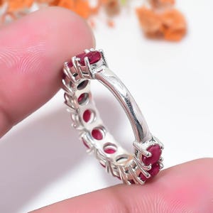 May include: A silver eternity band featuring a continuous row of round, deep red rubies set in individual metal prongs. The ring has a polished metallic finish and a uniform design that circles the entire circumference of the band.