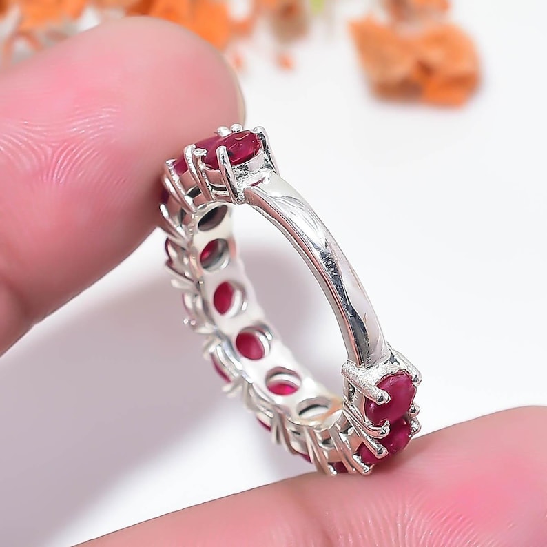 May include: A silver eternity band featuring a continuous row of round, deep red rubies set in individual metal prongs. The ring has a polished metallic finish and a uniform design that circles the entire circumference of the band.