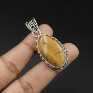 May include: An oval tiger's eye gemstone pendant set in a decorative silver frame. The stone features golden-brown tones with dark, chatoyant streaks. The silver bail at the top displays an intricate, openwork scroll design with small bead accents.