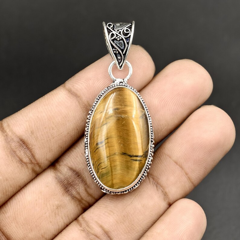 May include: An oval-shaped tiger's eye gemstone pendant set in a silver-toned metal frame. The stone features golden-brown bands with a chatoyant luster. The pendant includes a decorative silver-toned bail with intricate scrollwork detailing.