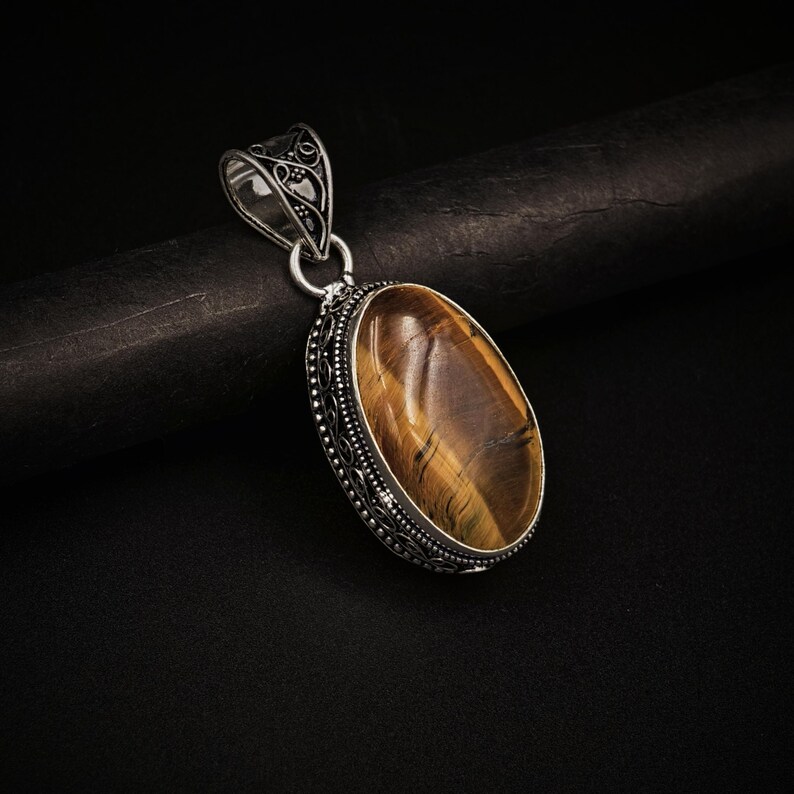 May include: An oval tiger's eye gemstone pendant set in a decorative silver frame with intricate metalwork detailing. The pendant features a polished, golden-brown stone with natural chatoyant bands and a matching silver bail at the top.