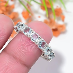 Puede incluir: Un anillo de banda de plata con una hilera de siete piedras preciosas redondas de color azul claro facetadas, engastadas en garras de metal individuales. Las piedras están dispuestas en una línea continua a lo largo de la parte superior del anillo.