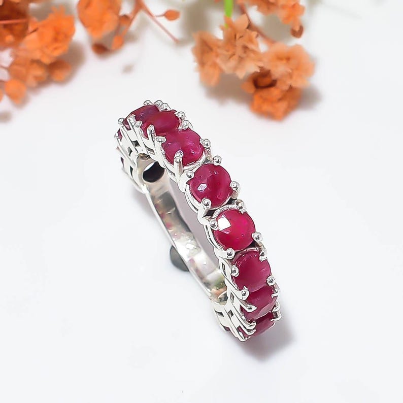 May include: A silver band ring featuring a row of seven round, deep red ruby gemstones set in individual prongs. The metal band has a polished finish, and the stones are arranged in a continuous line along the top half of the ring structure.