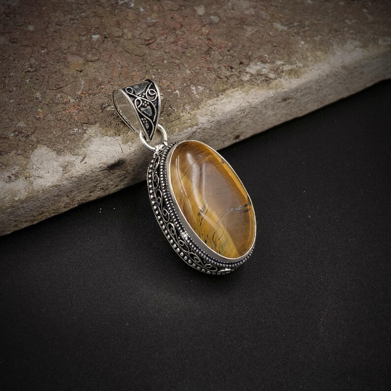 May include: An oval tiger's eye gemstone pendant set in a decorative sterling silver frame with intricate filigree detailing. The pendant features a polished, golden-brown stone with natural chatoyant bands and a matching silver bail at the top.