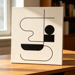 May include: Square canvas art with a minimalist black and white geometric design. The artwork features a black circle, rectangle, and curved lines on a cream background. The canvas is displayed on a wooden surface.