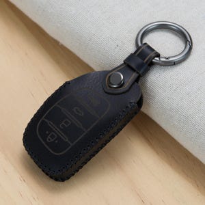 May include: A dark charcoal gray leather car key fob cover featuring a textured surface, visible stitched edges, a central metallic snap button, and a circular gunmetal keyring attachment. The cover displays embossed outlines of vehicle control icons.