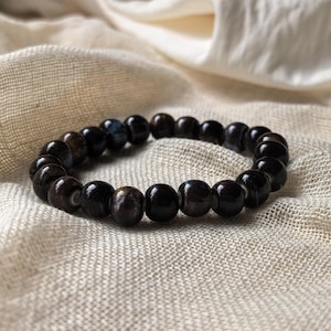 Könnte beinhalten: Ein dunkelbraunes Perlenarmband mit poliertem Finish. Das Armband besteht aus runden Perlen und liegt auf einem strukturierten, cremefarbenen Stoff. Die Perlen weisen subtile Farbvariationen mit Andeutungen von Blau und hellem Braun auf.