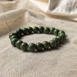 Könnte beinhalten: Ein grünes Perlenarmband. Das Armband besteht aus runden, dunkelgrünen Perlen. Die Perlen sind zu einer kreisförmigen Form aufgefädelt. Das Armband liegt auf einer beigen, strukturierten Oberfläche.