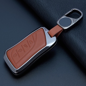 May include: A rectangular car key fob cover featuring a textured tan leather insert framed by a polished dark metallic border. The cover includes a matching tan leather strap connected to a dark metallic carabiner-style clip with a circular center.