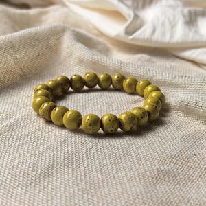 May include: A beaded bracelet featuring olive-green, speckled beads. The bracelet is circular and rests on a textured, off-white fabric. The beads have a matte finish and are evenly spaced around the elastic band.